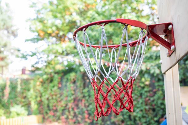 Red basketball hoop with white and red net against a wooden wall, green foliage in the background.