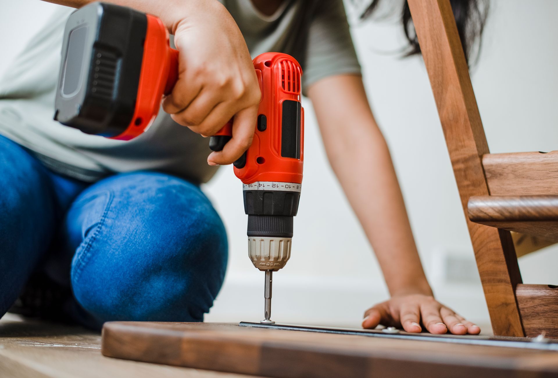 Person using a red and black power drill to assemble wooden furniture on a floor.