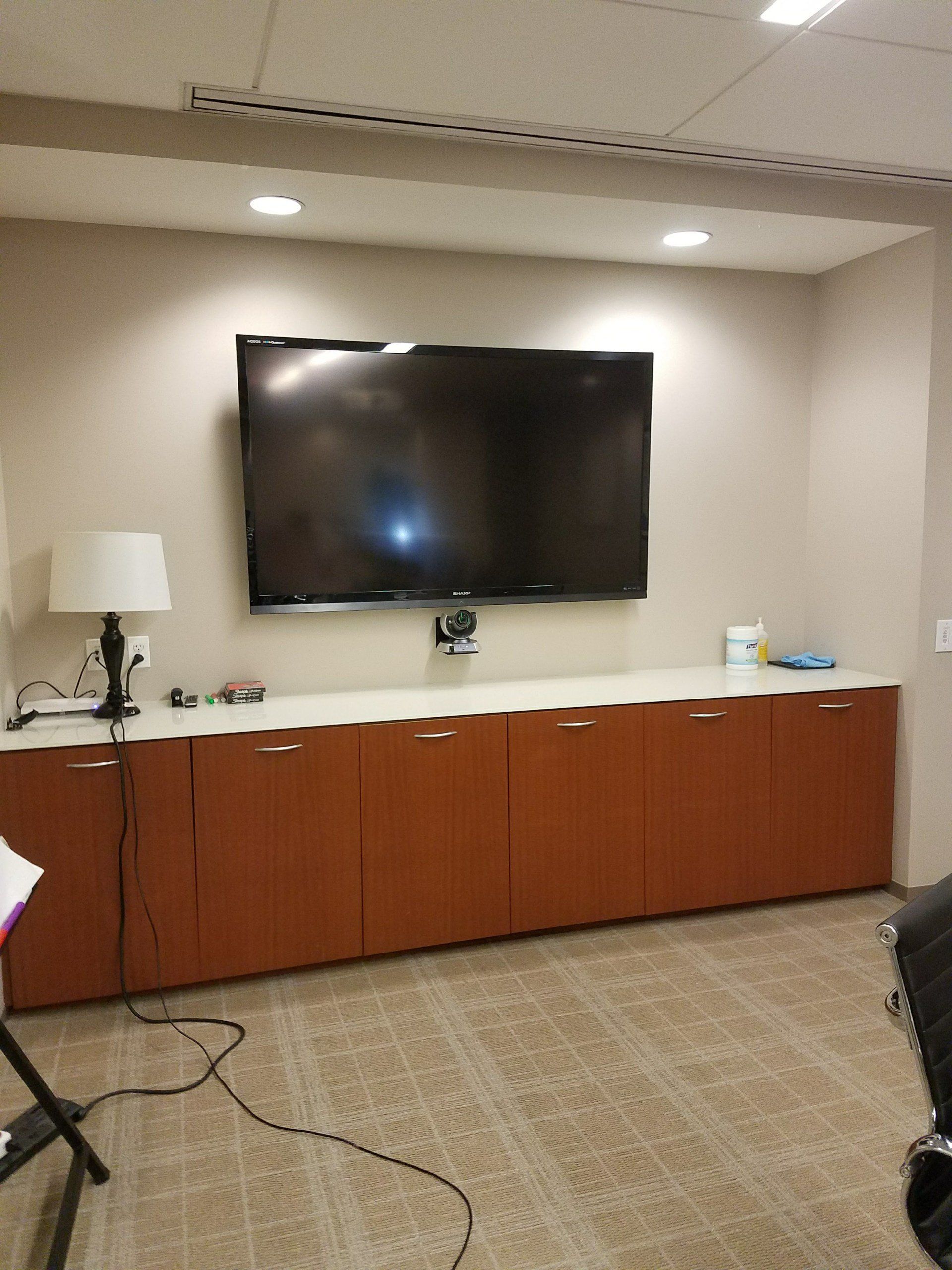 A large flat-screen TV mounted on a beige wall above a row of light brown wooden storage cabinets in an office space.