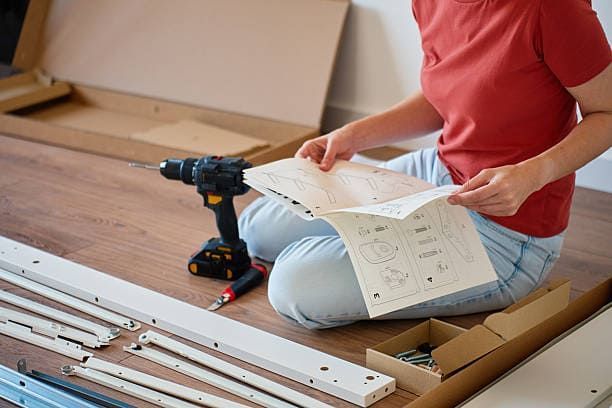 Person assembling furniture, reading instructions, surrounded by parts and tools.