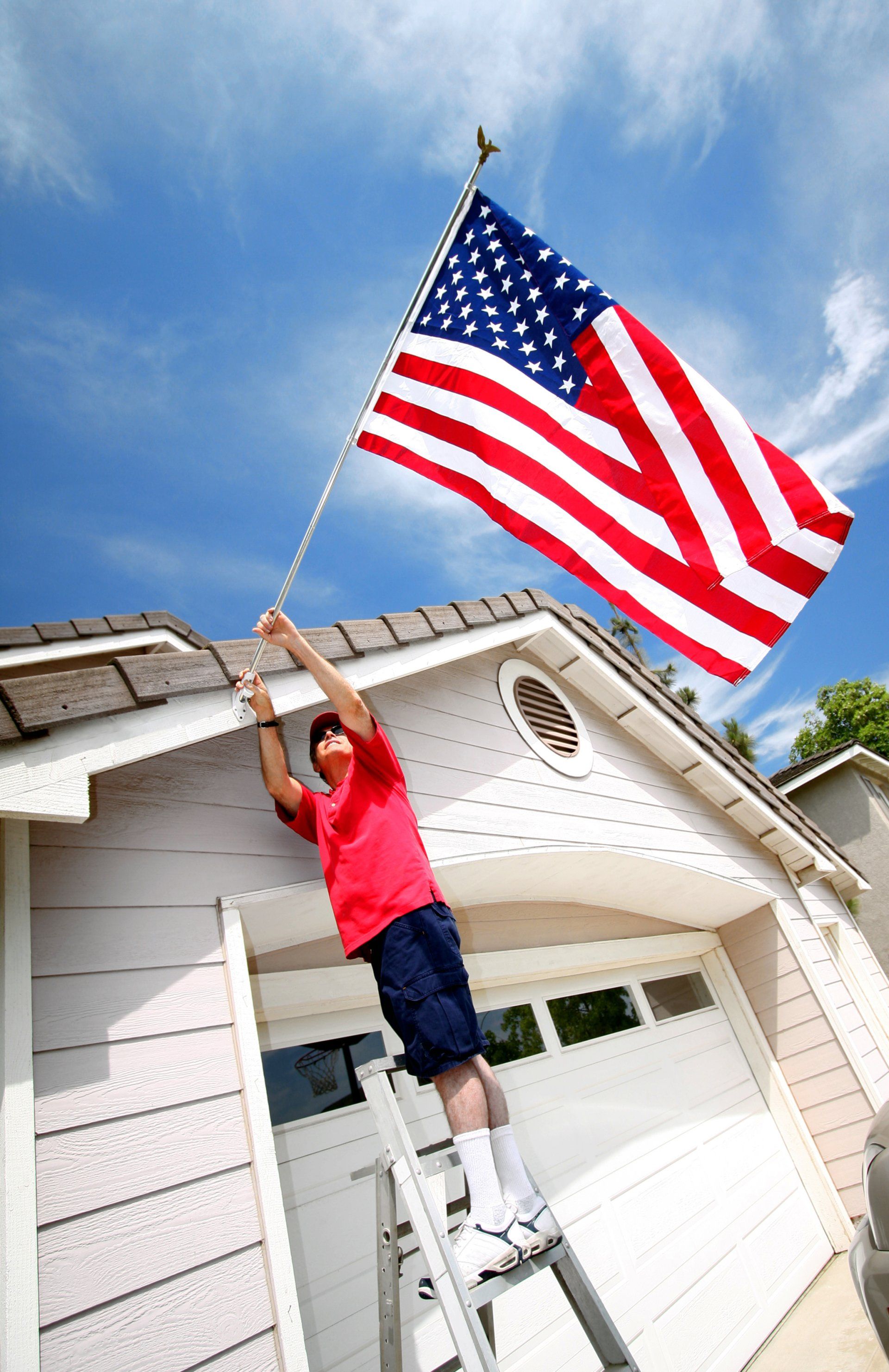 A man on a ladder is hanging an american flag on the roof of a house.