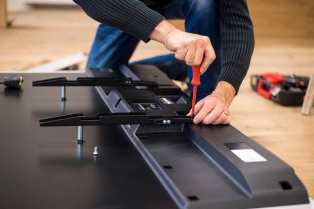 A person uses a red screwdriver to attach metal wall-mount brackets to the back of a black television.