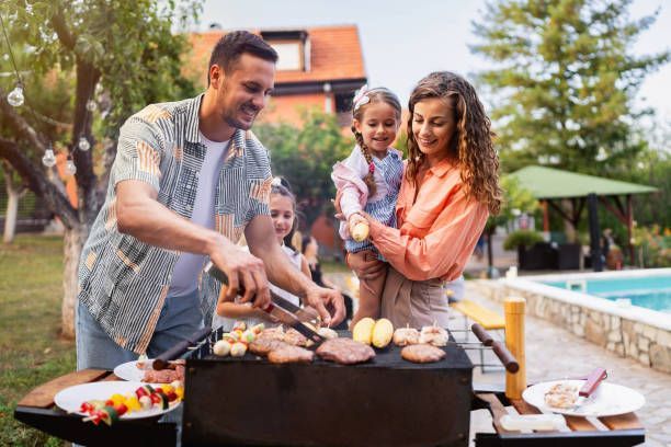 A family has a backyard barbecue, with an adult grilling food while another adult holds a child nearby near a pool.