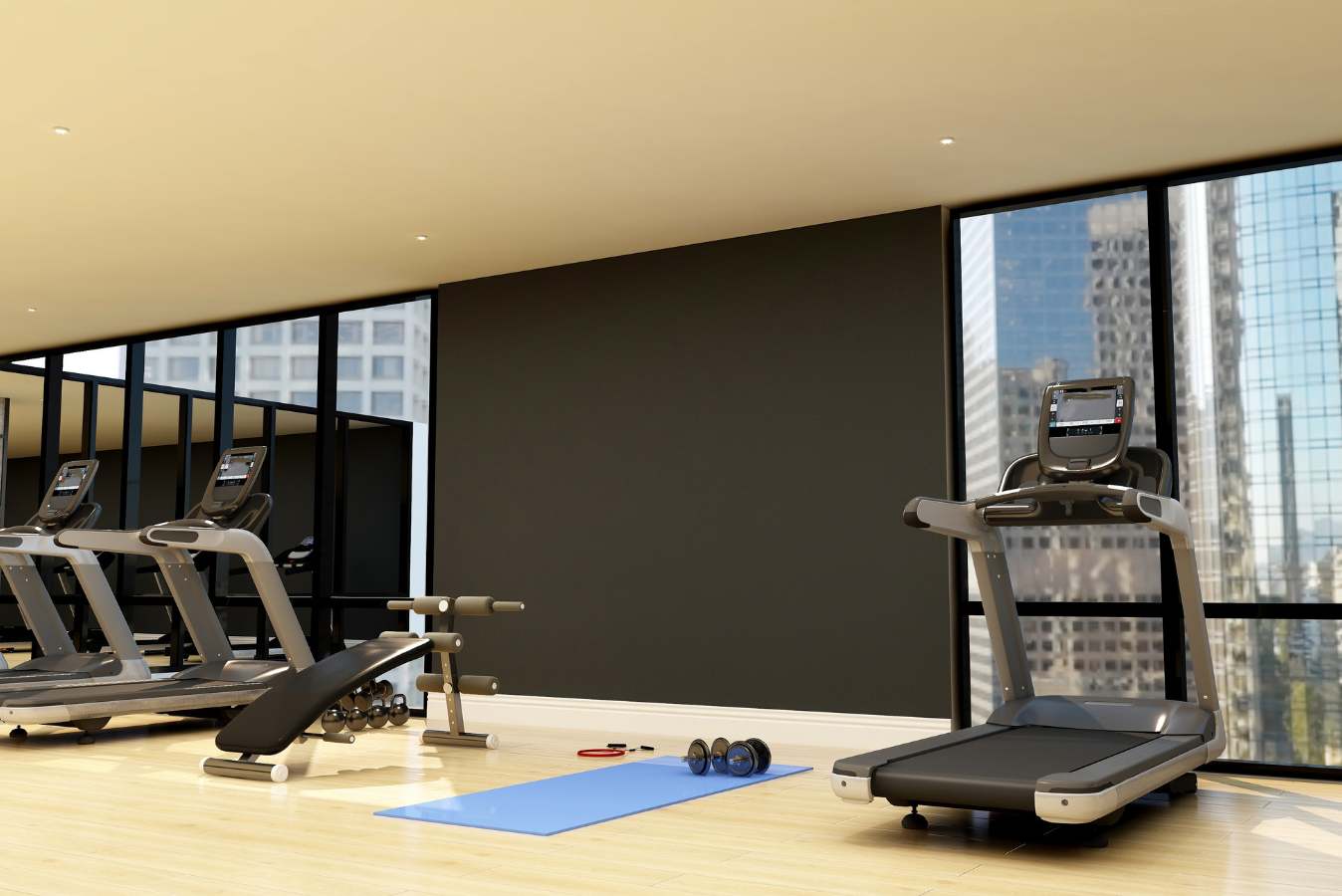 A modern gym with treadmills, a weight bench, a blue yoga mat with dumbbells, and large windows overlooking a city skyline.