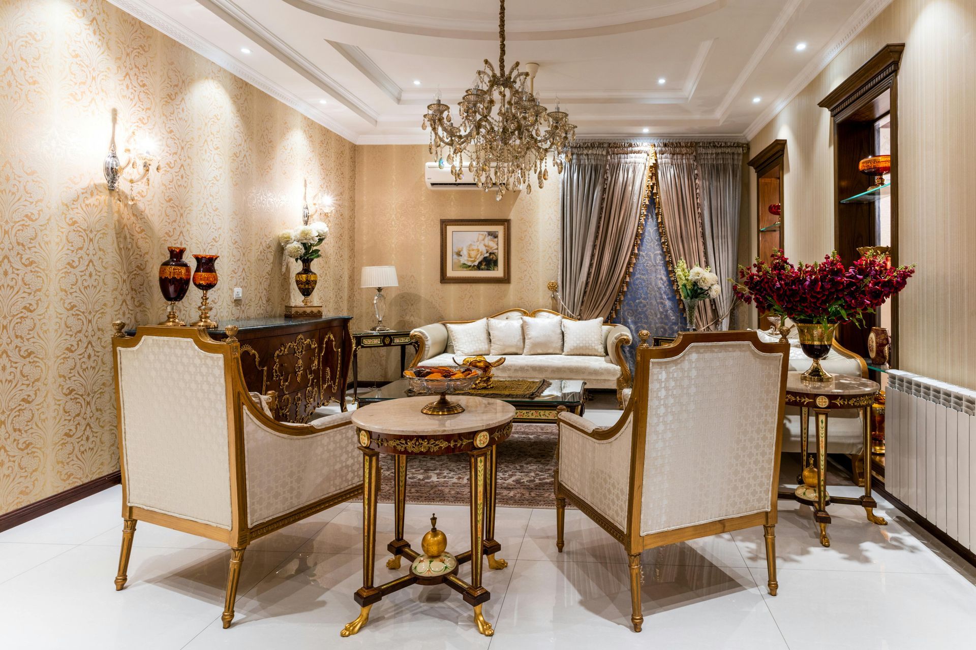 Elegant beige and gold living room with ornate chairs, a sofa, chandelier, and floral arrangements.