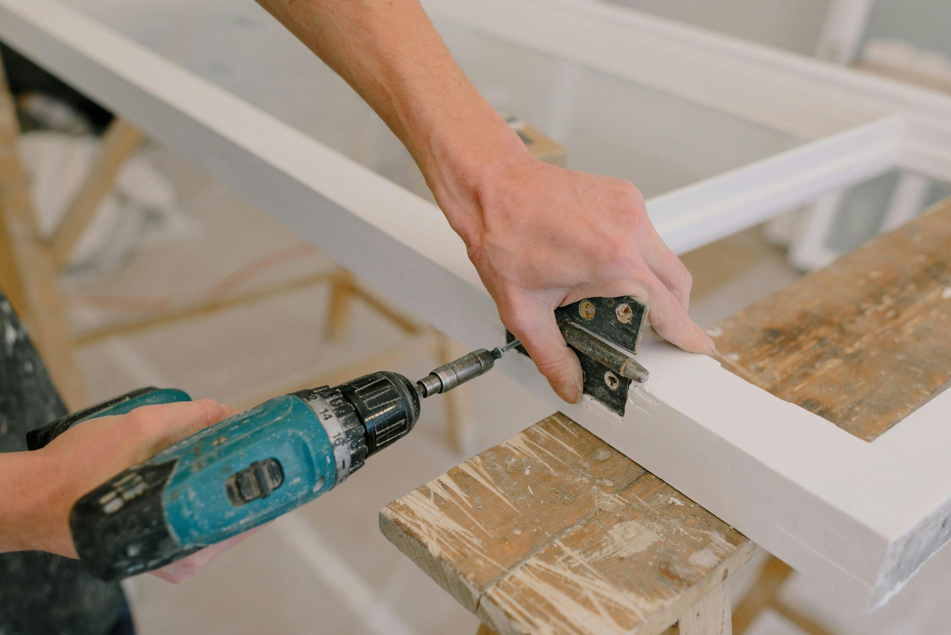 A person uses a cordless drill to attach a metal hinge to a white door frame resting on a wooden sawhorse.
