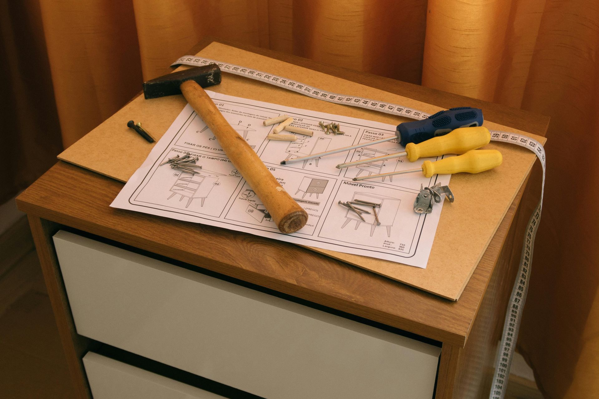 A hammer, screwdrivers, hardware, and assembly instructions on a partially assembled wooden bedside table.