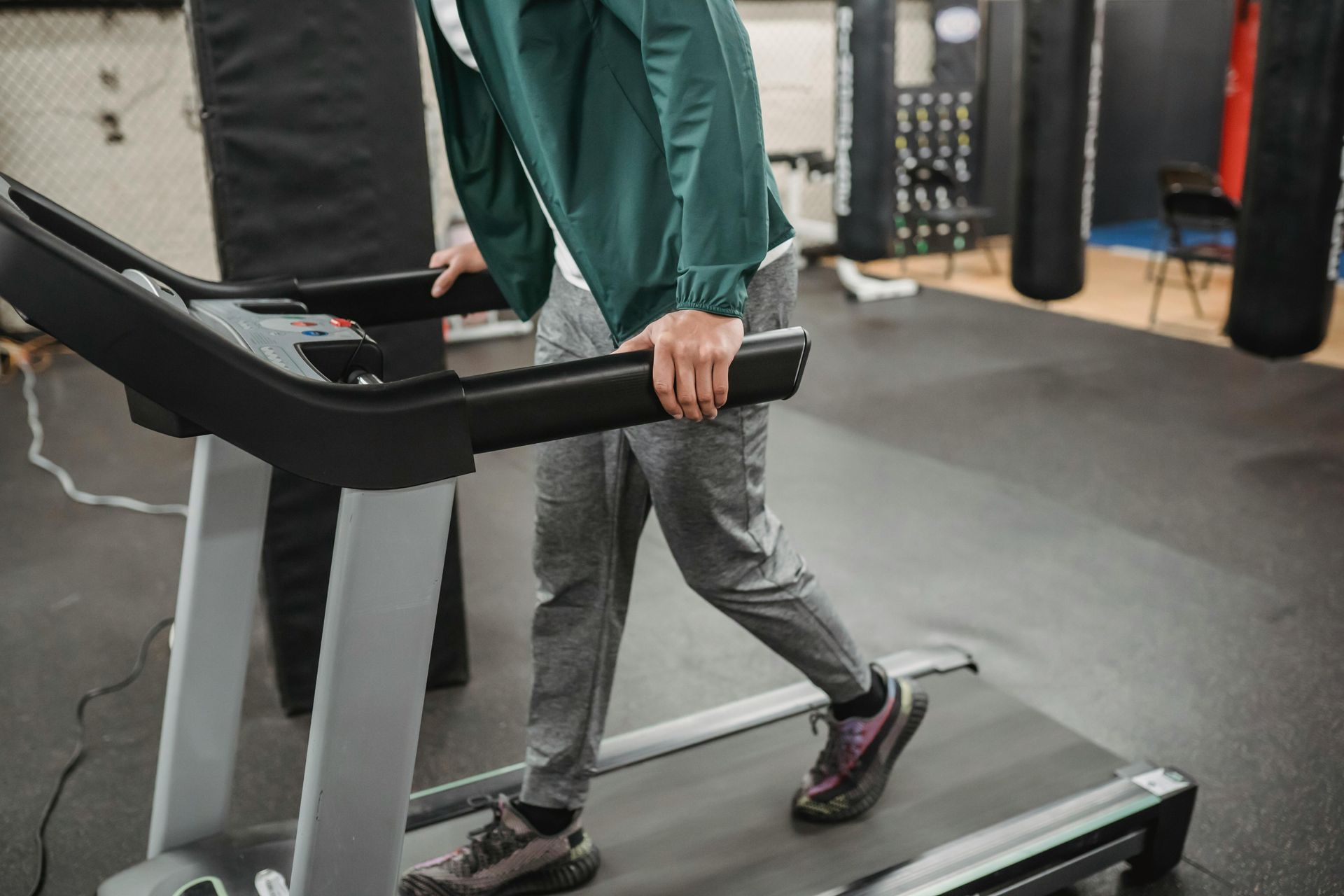 Person walking on a treadmill in a gym, holding the rails and wearing a green jacket and gray pants