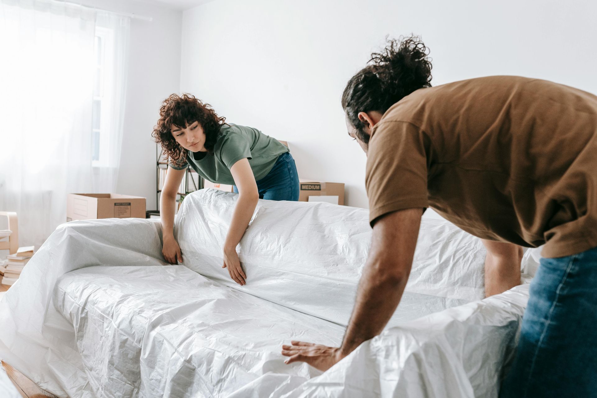 Two people covering a couch with a white sheet in a room with cardboard boxes.