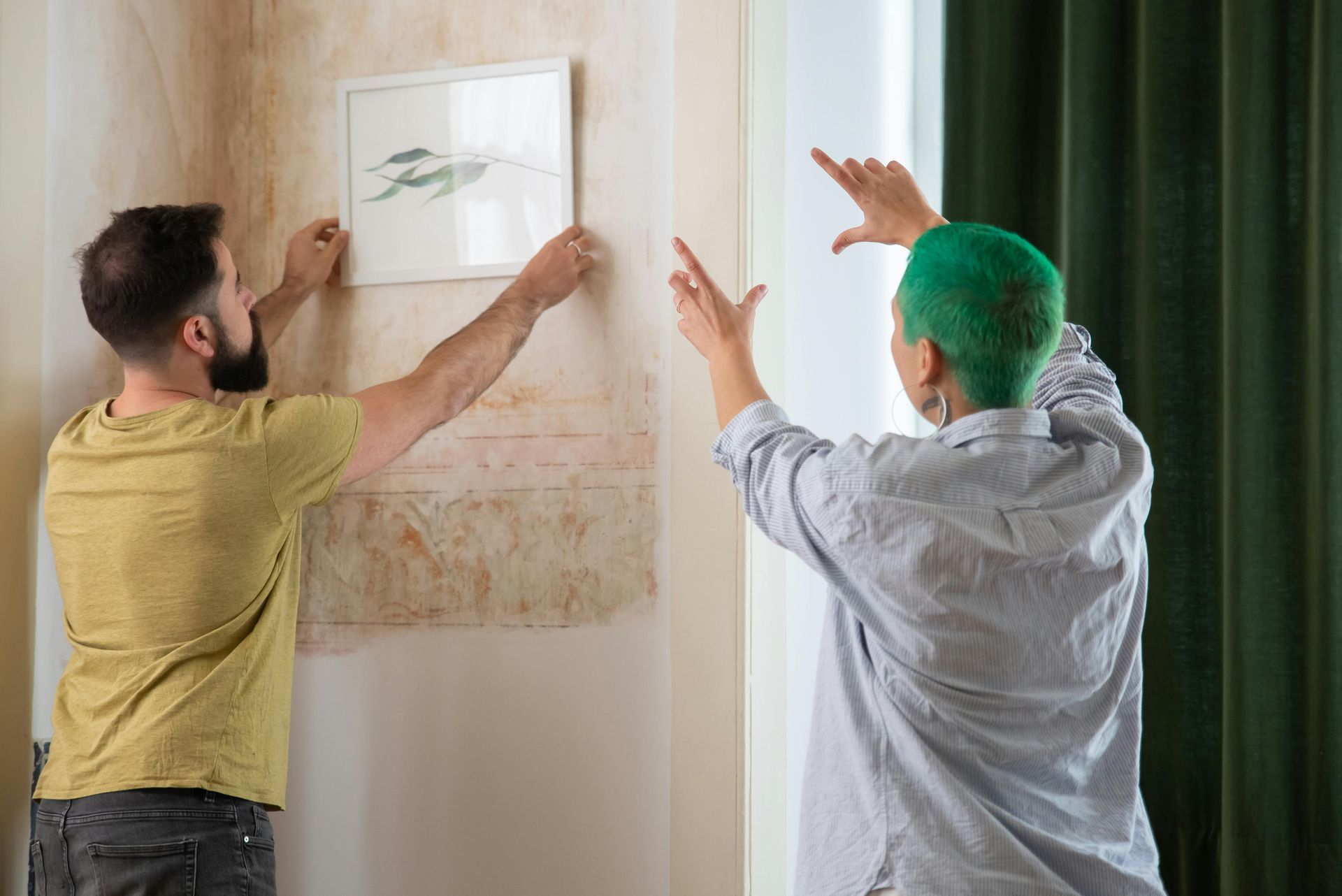 Two people hanging a framed picture on a wall. One is holding it, the other is gesturing.