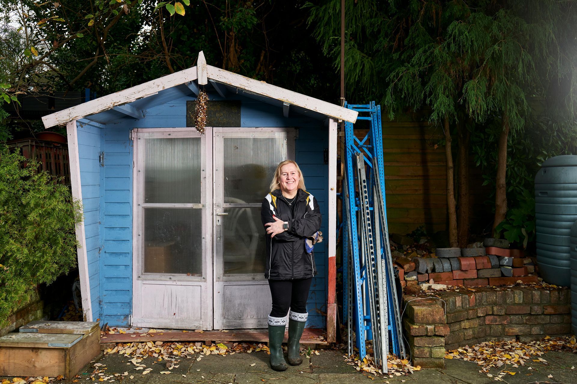 Woman standing in front of a blue shed, arms crossed. She wears a black jacket and green boots.
