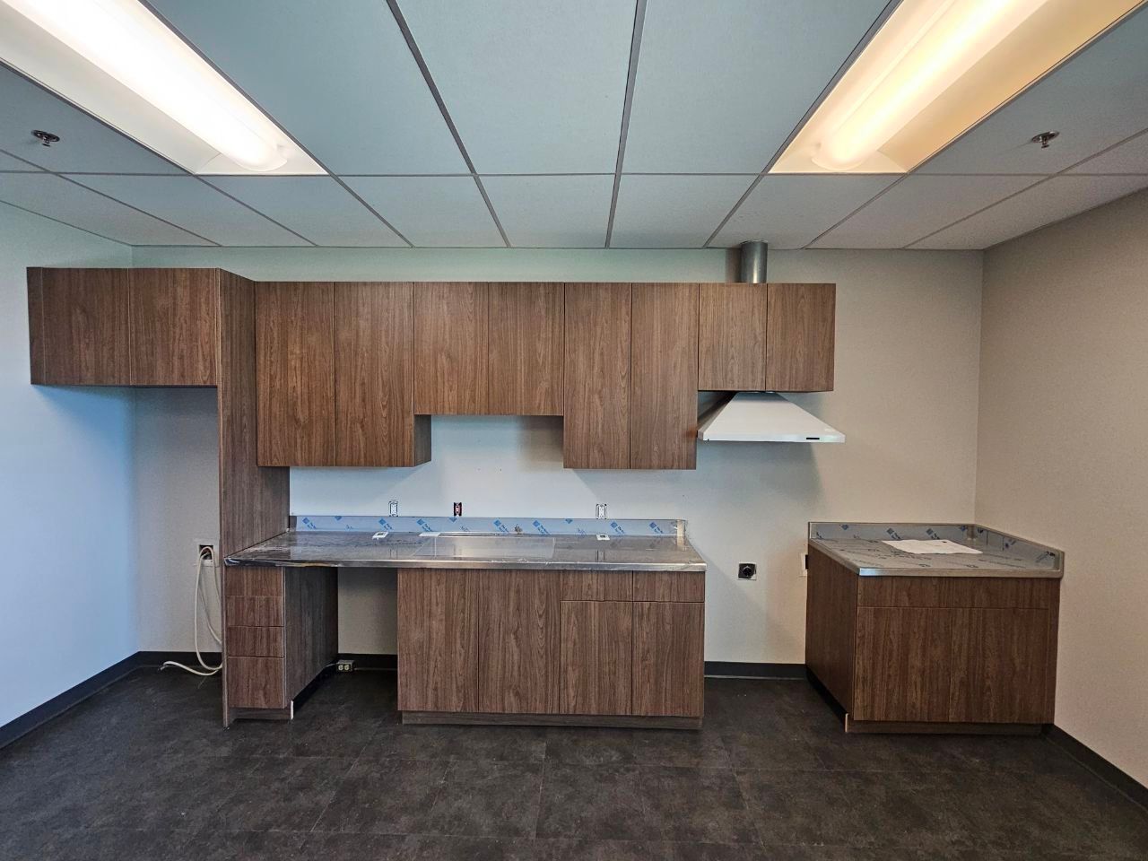 A breakroom with brown wood cabinets, metal countertops, a range hood, and dark flooring in a drop-ceiling office space.