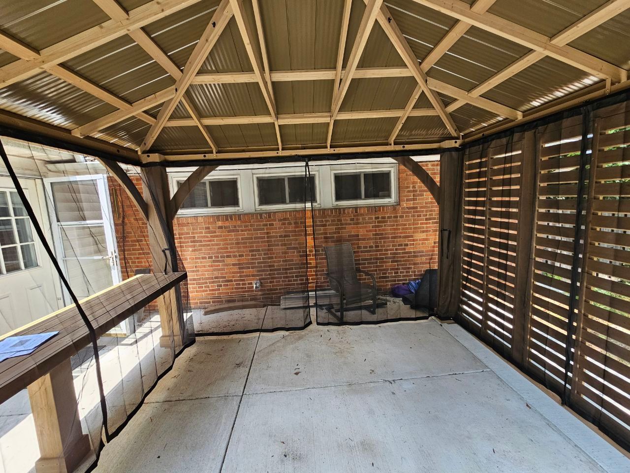 Gazebo interior with concrete floor, wooden frame, and brick wall backdrop.
