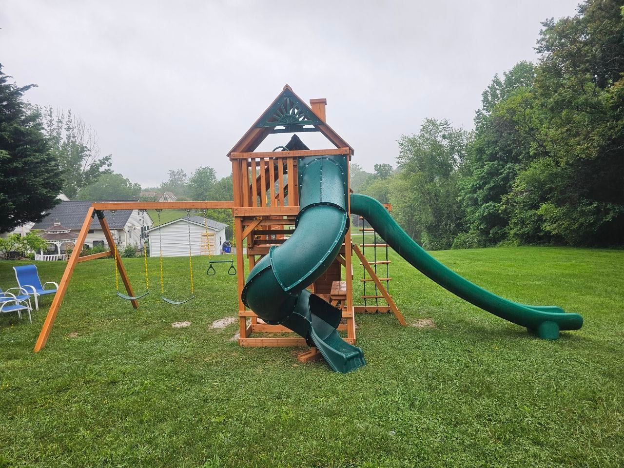 A wooden playground structure with a green tube slide, a straight green slide, and a swing set in a grassy yard.