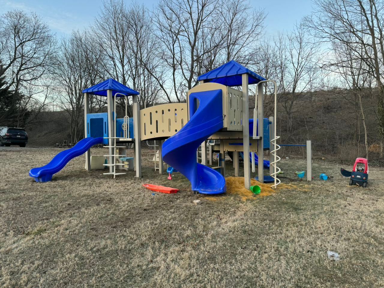 Playground with blue slides, beige climbing wall, and brown grass. Trees in the background.