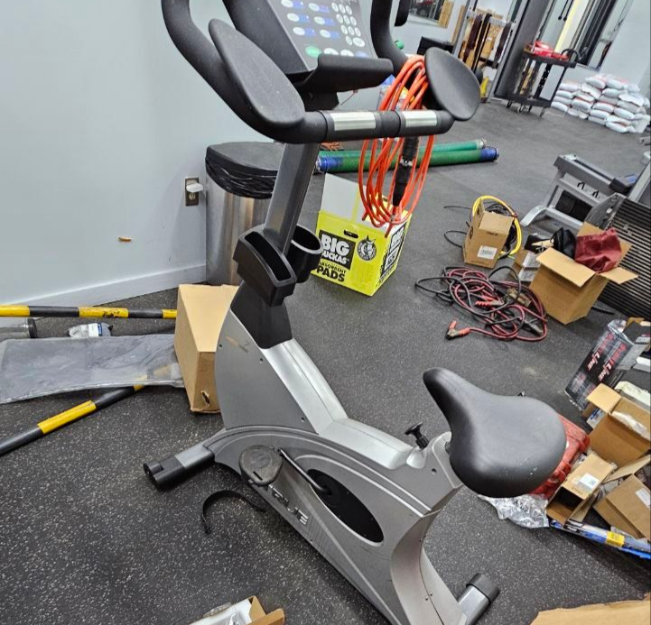 A silver stationary exercise bike sits in a cluttered gym area with cardboard boxes and workout equipment nearby.