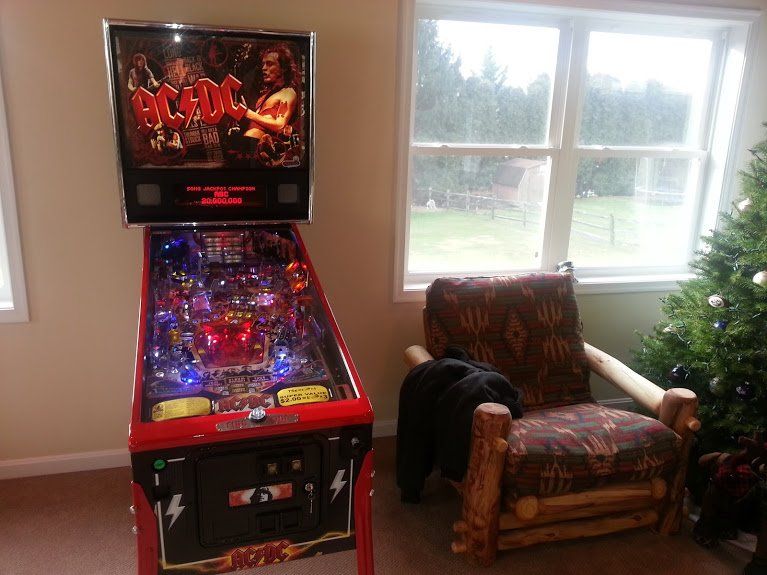 A pinball machine is sitting in a living room next to a chair and a christmas tree.