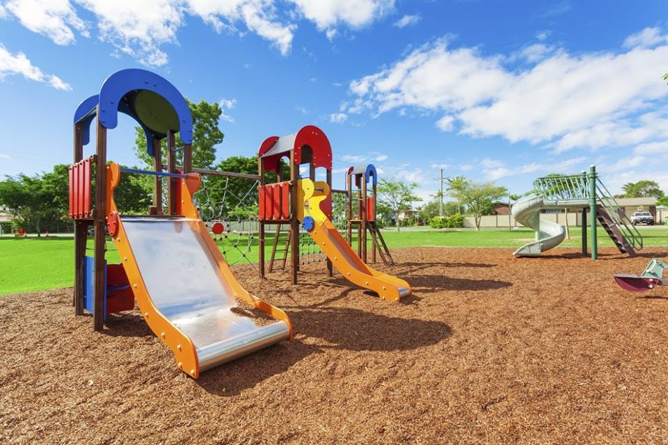 A playground with colorful climbing structures and slides on a wood-chip surface under a blue sky.