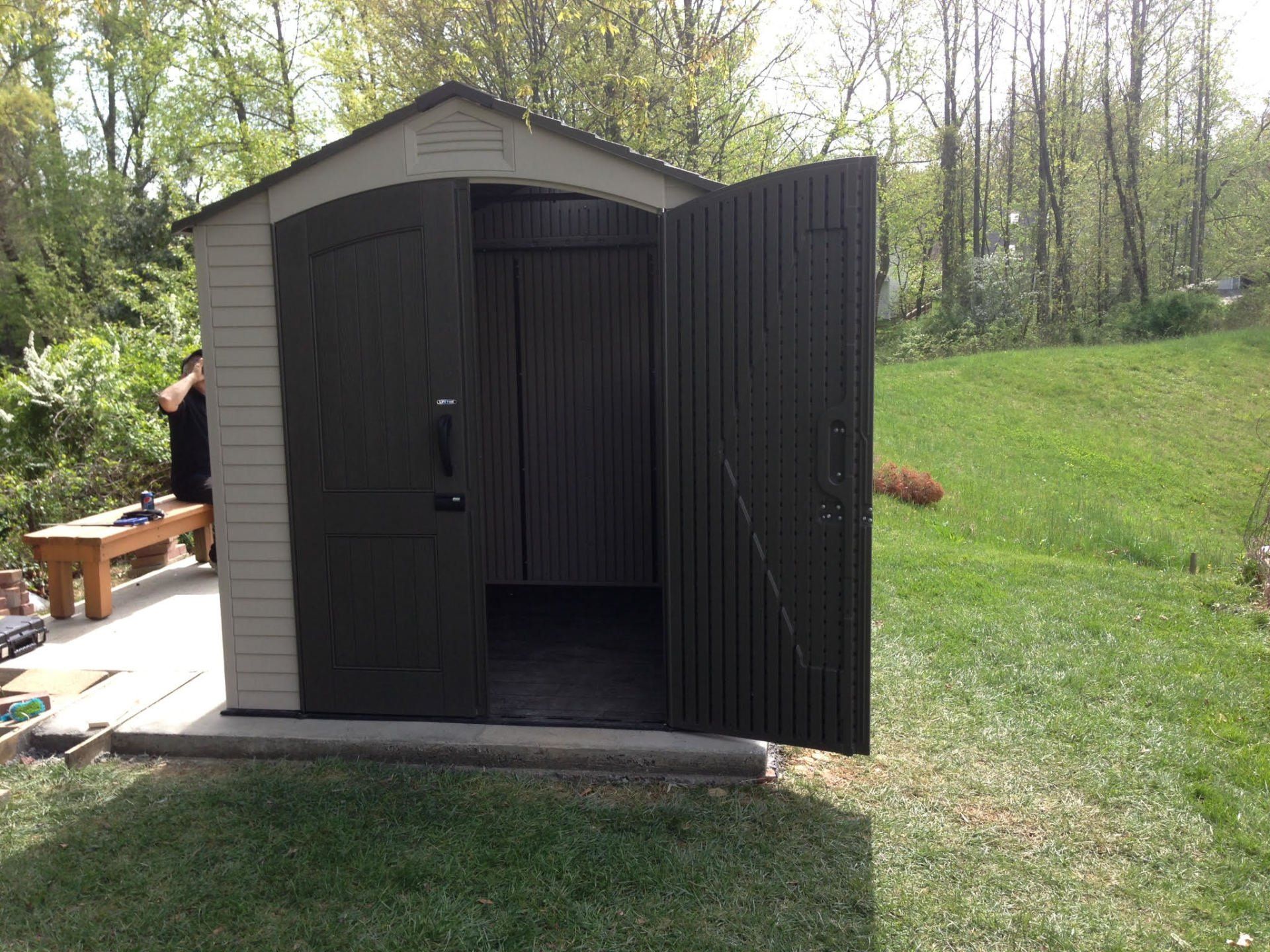 A man is sitting on a bench in front of a shed with its doors open.