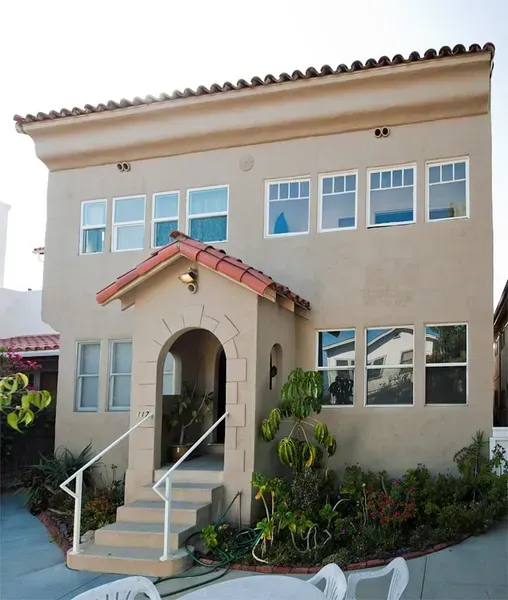Two-story beige stucco house with red-tiled porch roof, white-trimmed windows, and front steps