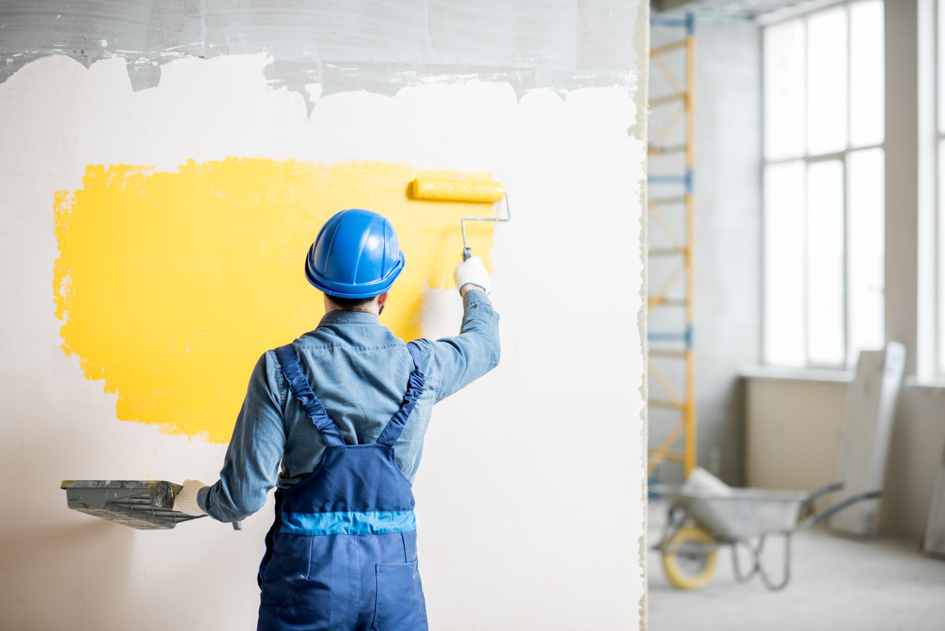 Painter in blue overalls applying bright yellow paint to a white wall in a room under renovation