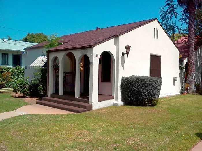 Small white stucco house with arched porch, red roof, and front lawn in sunny daylight