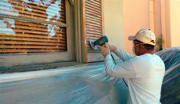 Worker spraying paint on a window frame and wall beside a shuttered opening.
