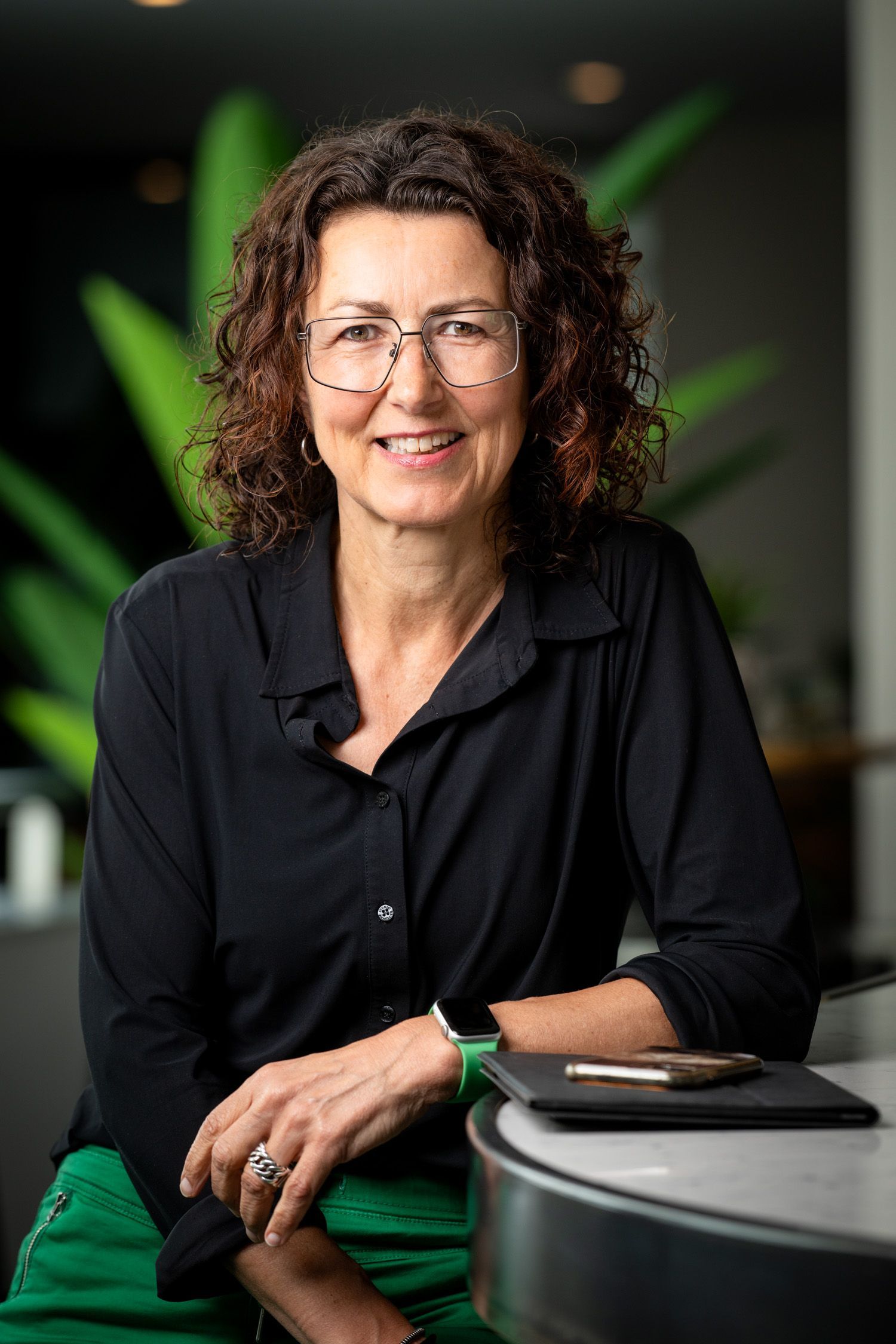 Een vrouw in een zwarte blouse glimlacht naast een tafel met een groene stoel in een vaag wazige kantooromgeving.