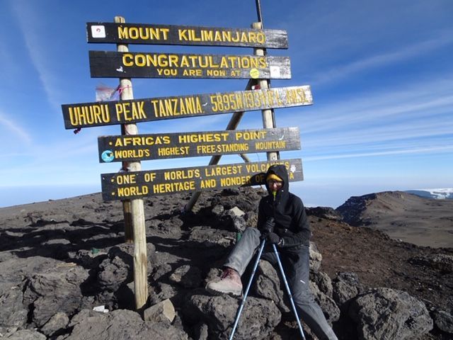 Een bord op de top van de Kilimanjaro met een wandelaar ernaast, op rotsachtig terrein onder een blauwe hemel.