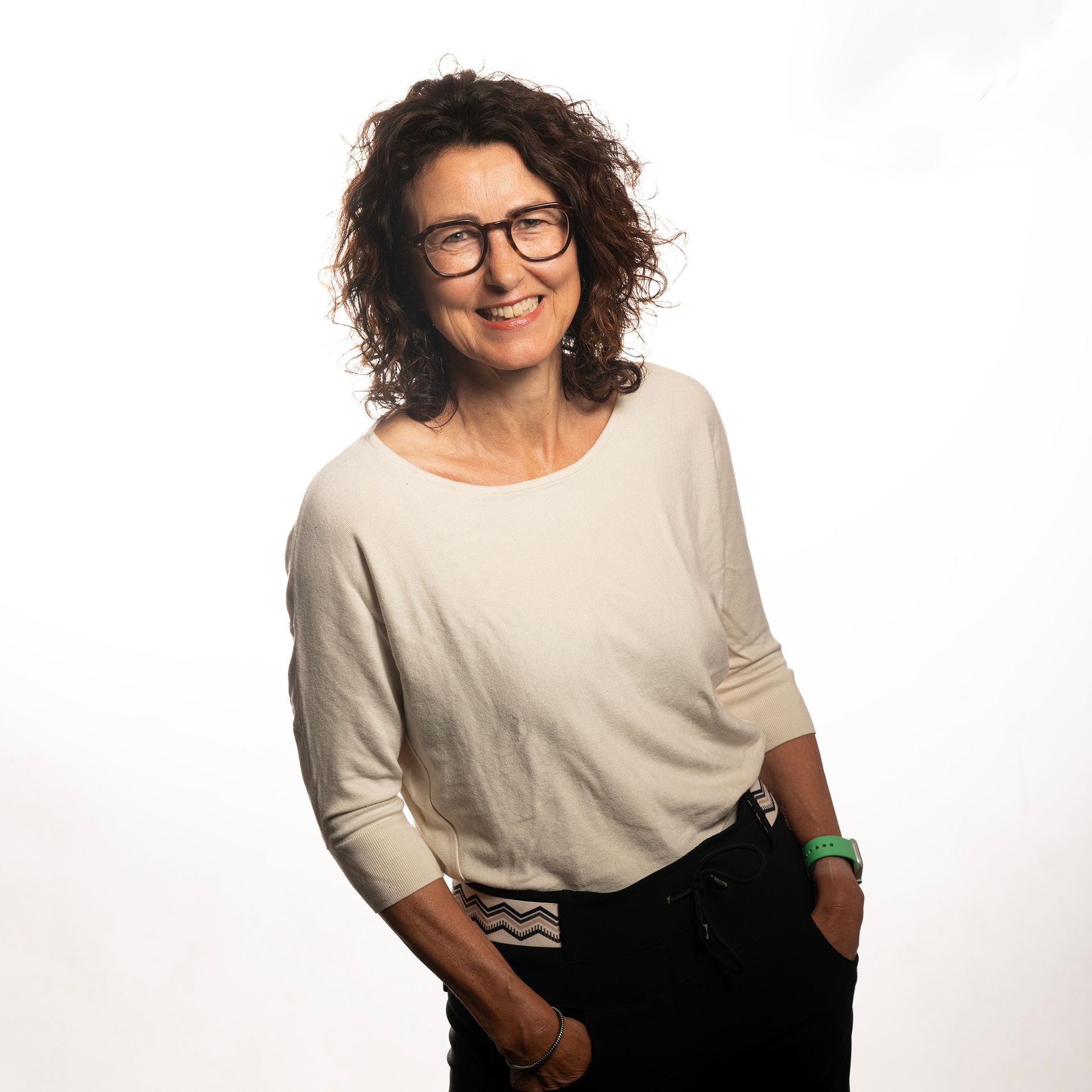 Een lachend persoon met krullend haar en een bril, gekleed in een crèmekleurig topje, tegen een witte achtergrond.