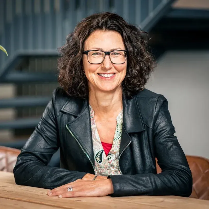 Een glimlachend persoon met een bril en een zwarte leren jas zit aan een tafel met een trap op de achtergrond.