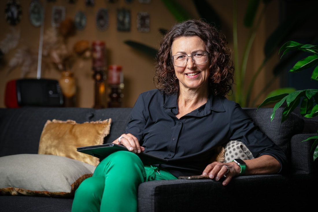 Vrouw zit op een bank in een gezellige woonkamer, glimlachend met een kat achter haar.