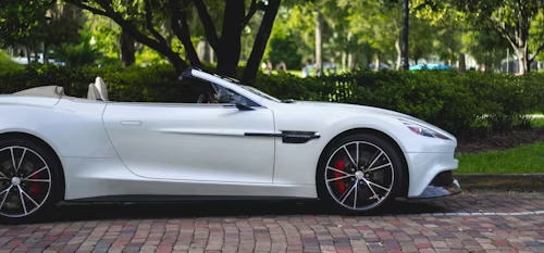 White convertible Aston Martin parked on a brick-paved surface, with trees and green hedges in the background.
