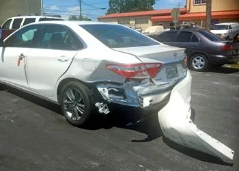 A white sedan parked in a lot with its rear bumper detached and dragging on the ground due to collision damage.