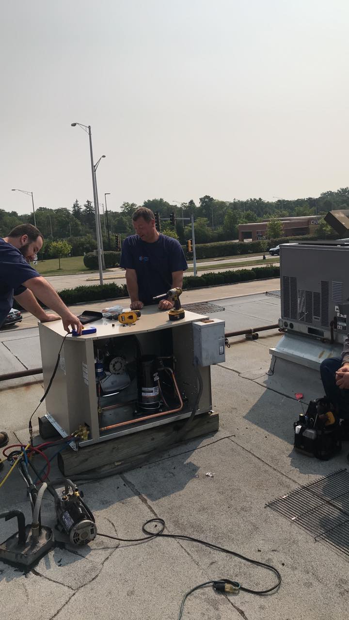 Two men are working on an air conditioner on the roof of a building.