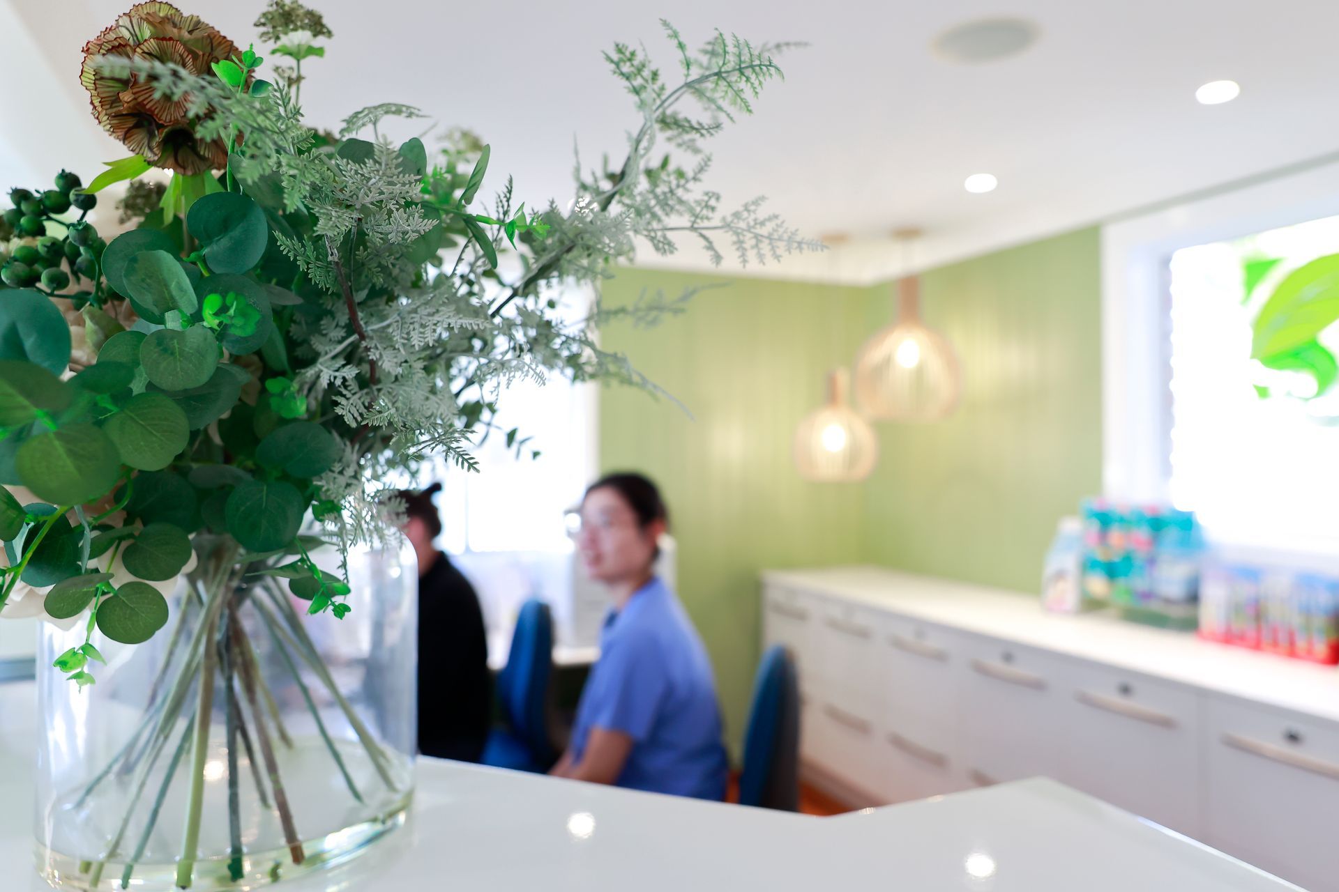 Vase of greenery on a white counter; blurred people and a white desk in the background. Green wall - Reception area O'Reilly Dental