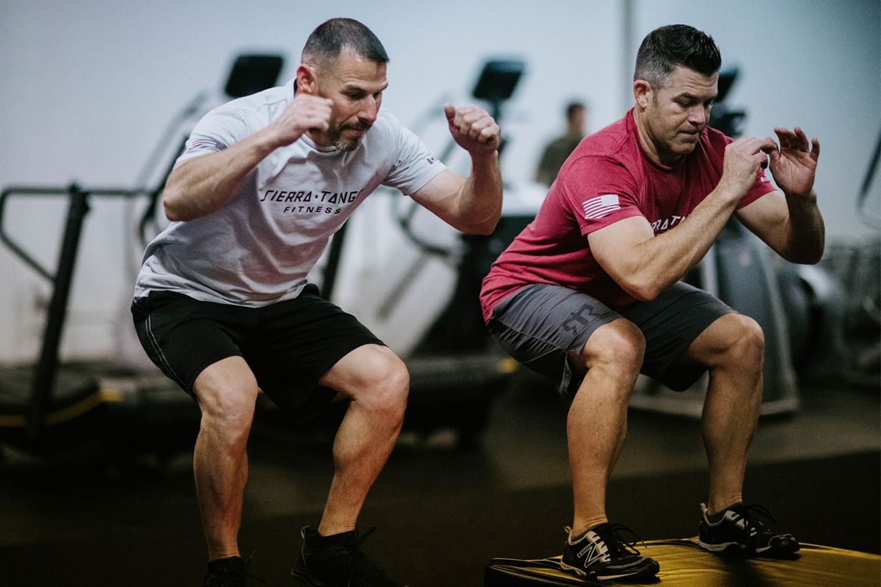 Two men are squatting on a platform in a gym.