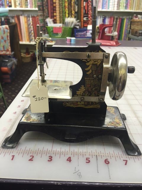 Antique sewing machine with black base and silver details, priced at $230, on a cutting mat in a fabric store.