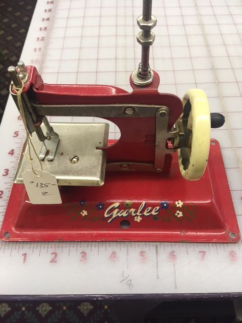 Red and cream Garlee toy sewing machine on a white grid surface.