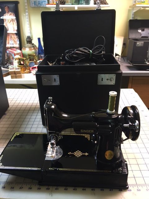 Black Singer sewing machine with open case, spool of green thread. Sitting on a cutting mat.