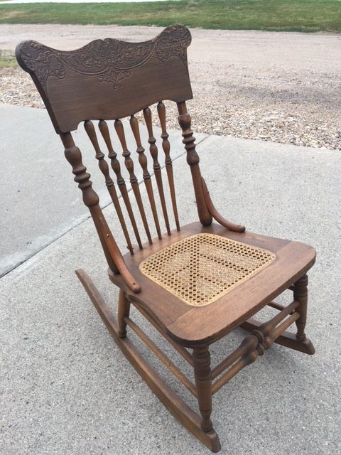 Wooden rocking chair with a woven seat and carved back, outdoors on concrete.