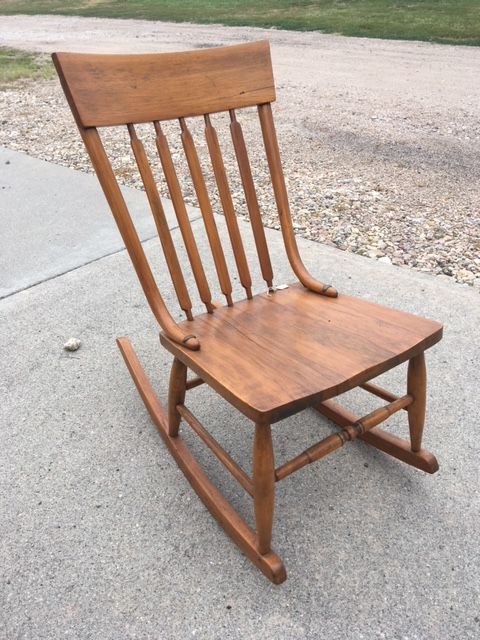 Wooden rocking chair on a concrete surface, with vertical back slats, a curved back, and a warm brown finish.