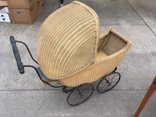 Wicker doll stroller with a woven hood and metal wheels, outdoors on concrete.