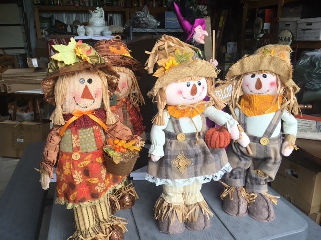 Three fall scarecrow dolls with straw legs and smiling faces, set in a garage.