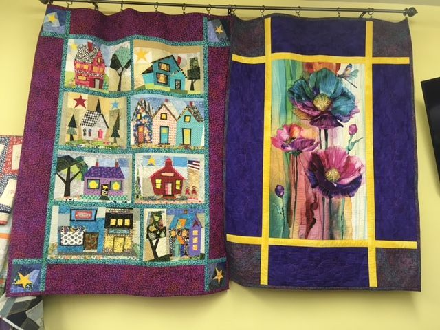 Two quilts: one with colorful houses, the other with watercolor flowers, on a yellow wall.