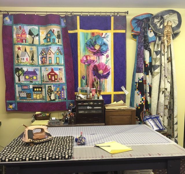 Quilting studio with table, iron, cutting mat, quilts on the wall, and sewing supplies.