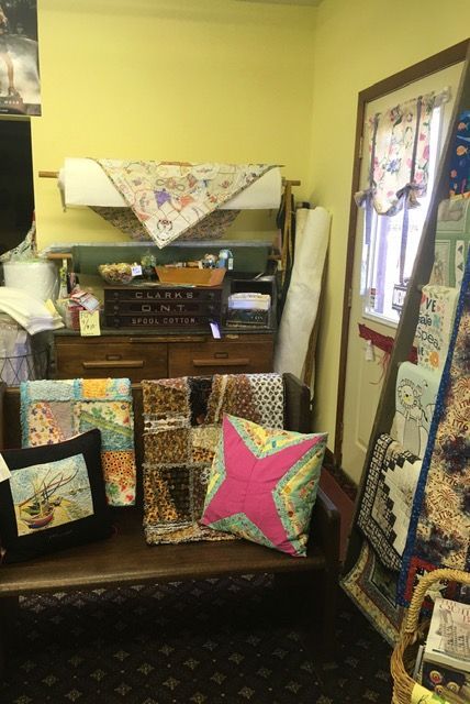 A craft shop corner with fabric rolls, quilts, pillows, and a wooden bench. Light yellow walls.