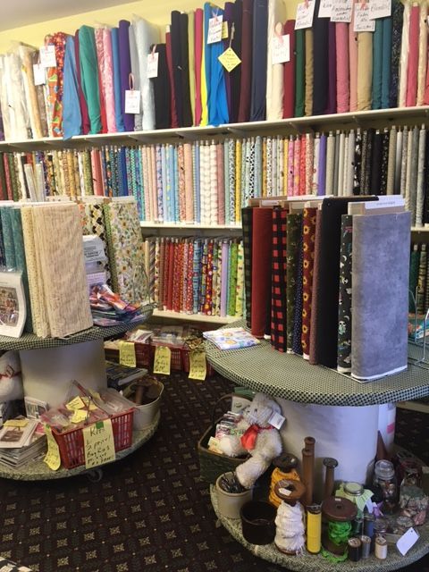 A fabric store interior with shelves stocked with colorful fabric bolts, plus round display tables with small items.