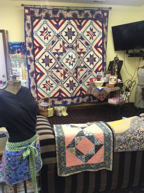 Quilts on display in a store, including a large red, white, and blue star quilt, a pink and blue quilt, and a mannequin.