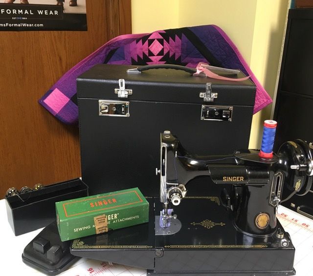 A black vintage Singer sewing machine with a matching case and a green Singer accessories box on a desk.